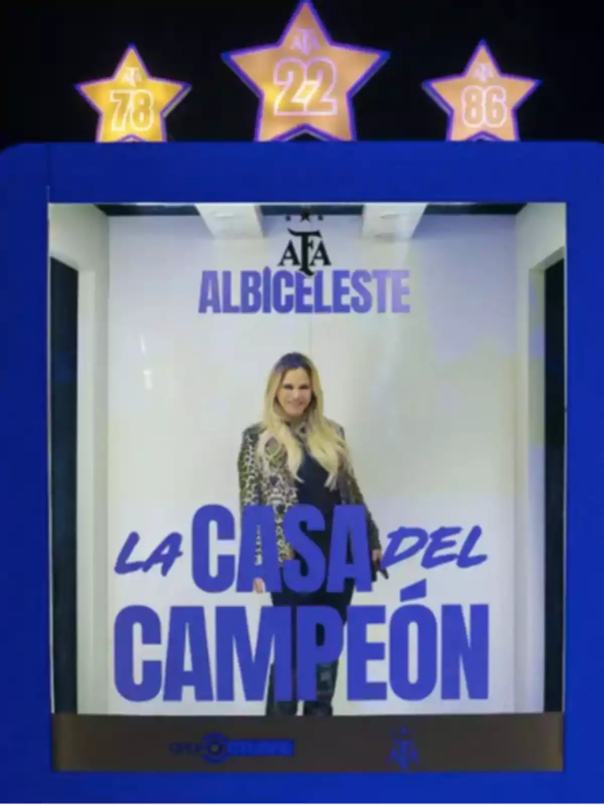 Una mujer rubia con saco animal print posando dentro de una cabina azul decorada con estrellas que tienen los números 78, 22 y 86, y el texto La casa del campeón junto al logo de la AFA. Una mujer rubia con saco animal print posando dentro de una cabina azul decorada con estrellas que tienen los números 78, 22 y 86, y el texto La casa del campeón junto al logo de la AFA.