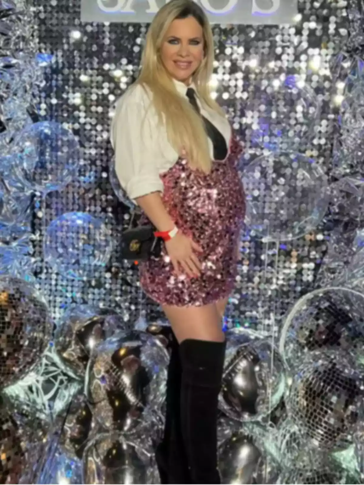Mujer rubia embarazada con vestido rosa brillante, camisa blanca y botas negras posando frente a una decoración de esferas y fondo plateado Mujer rubia embarazada con vestido rosa brillante, camisa blanca y botas negras posando frente a una decoración de esferas y fondo plateado