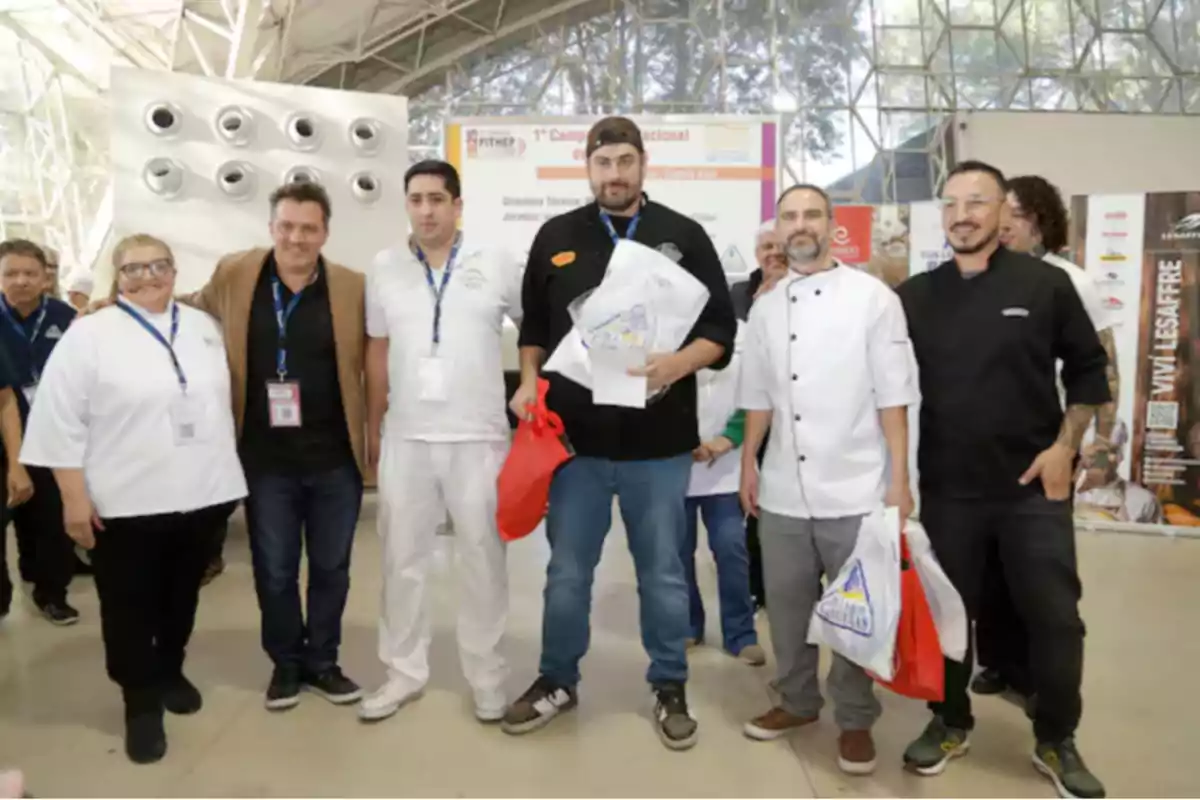 Group of people posing together at an event, some wearing chef uniforms and others casual clothing, holding bags and badges, in an indoor space with natural light.