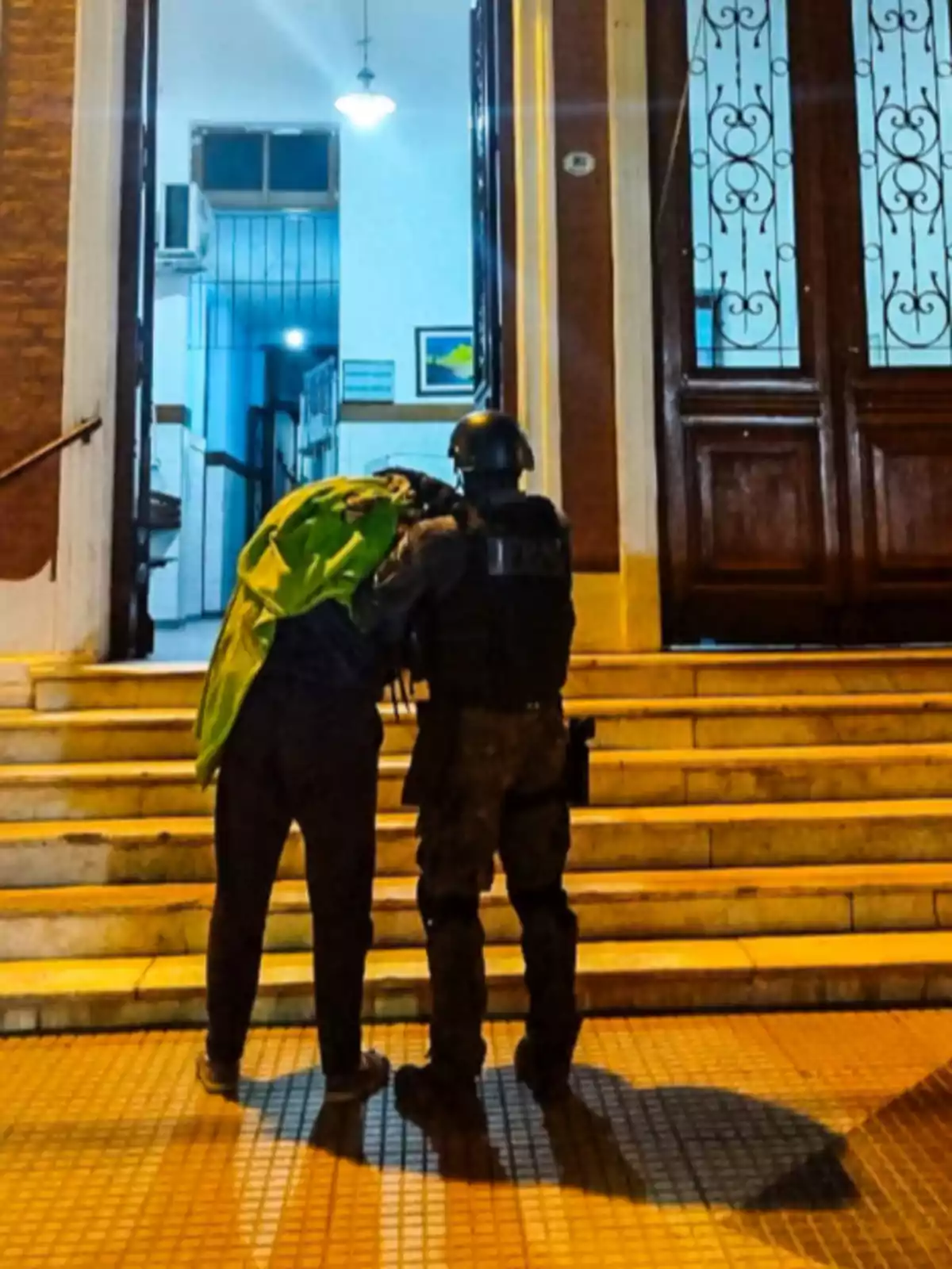 A police officer escorts a person with a green backpack as they go up the stairs of a building lit up at night. A police officer escorts a person with a green backpack as they go up the stairs of a building lit up at night.