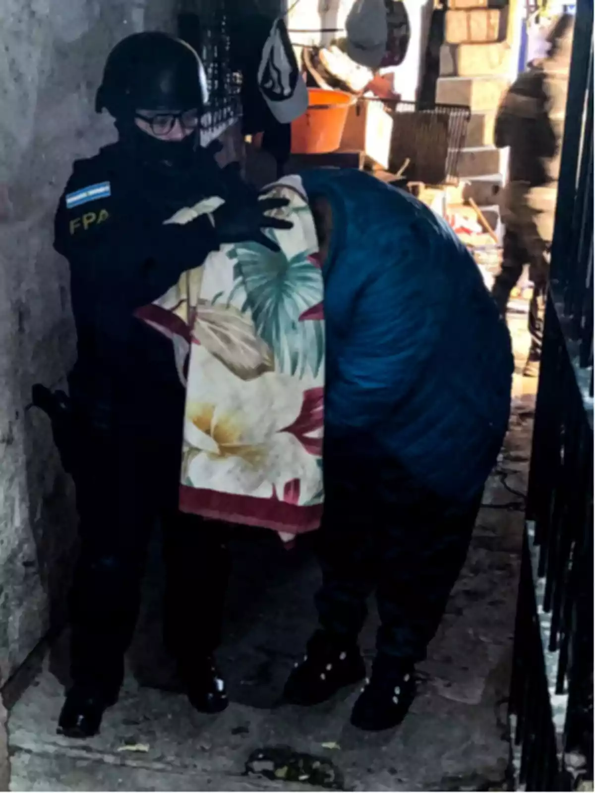 Policía con uniforme y casco escolta a una persona cubierta con una manta en un pasillo estrecho Policía con uniforme y casco escolta a una persona cubierta con una manta en un pasillo estrecho