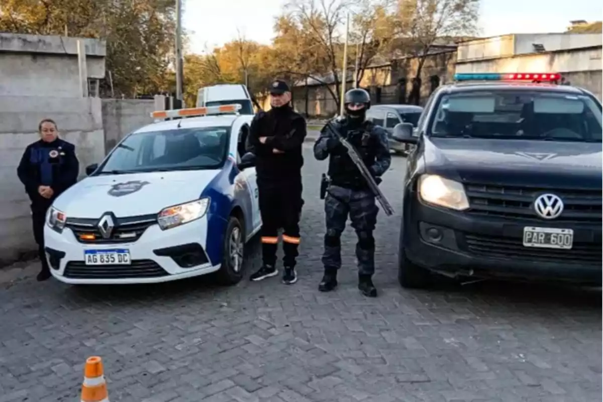 Tres policías de pie junto a dos patrullas en una calle, uno de ellos con uniforme táctico y un arma, mientras que los otros dos llevan uniforme estándar