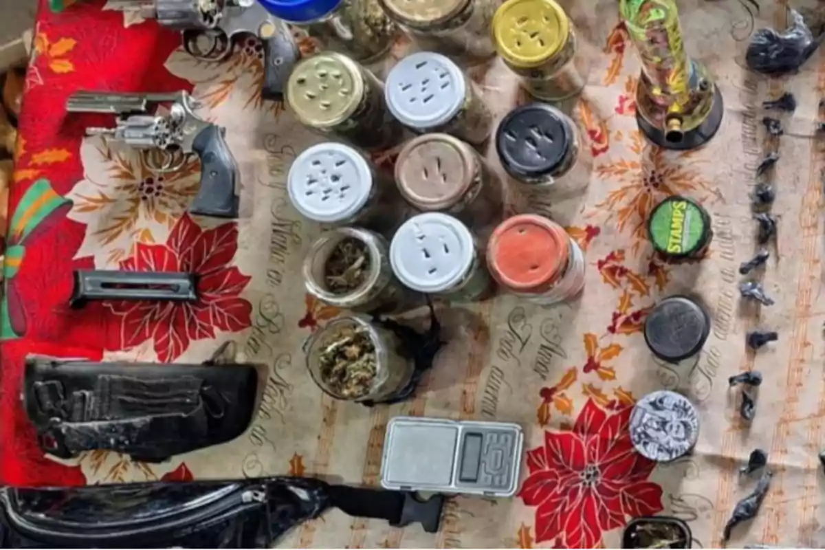 Objects related to weapons, jars with weed, a scale, a grinder, a bong, and small wrappers on a table with a red flower tablecloth.