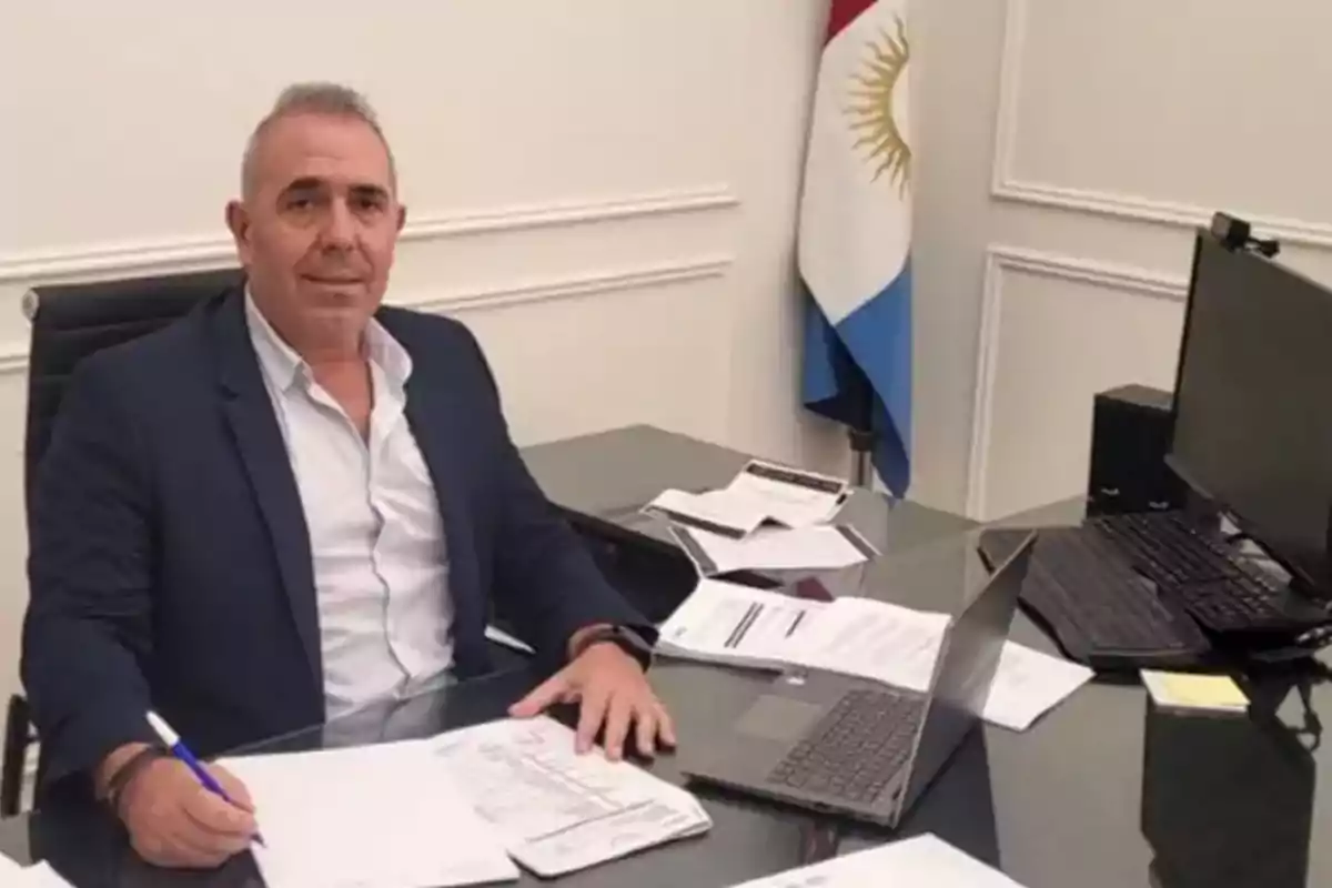 Hombre de traje sentado en un escritorio con papeles y una computadora portátil, al fondo una bandera de Argentina Hombre de traje sentado en un escritorio con papeles y una computadora portátil, al fondo una bandera de Argentina