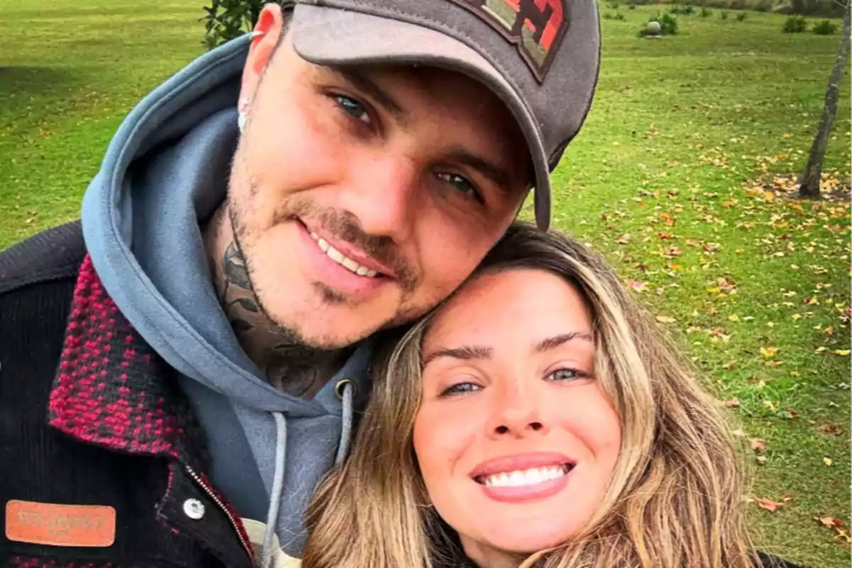 Young couple smiles while taking a selfie outdoors in a park with grass and fallen leaves.