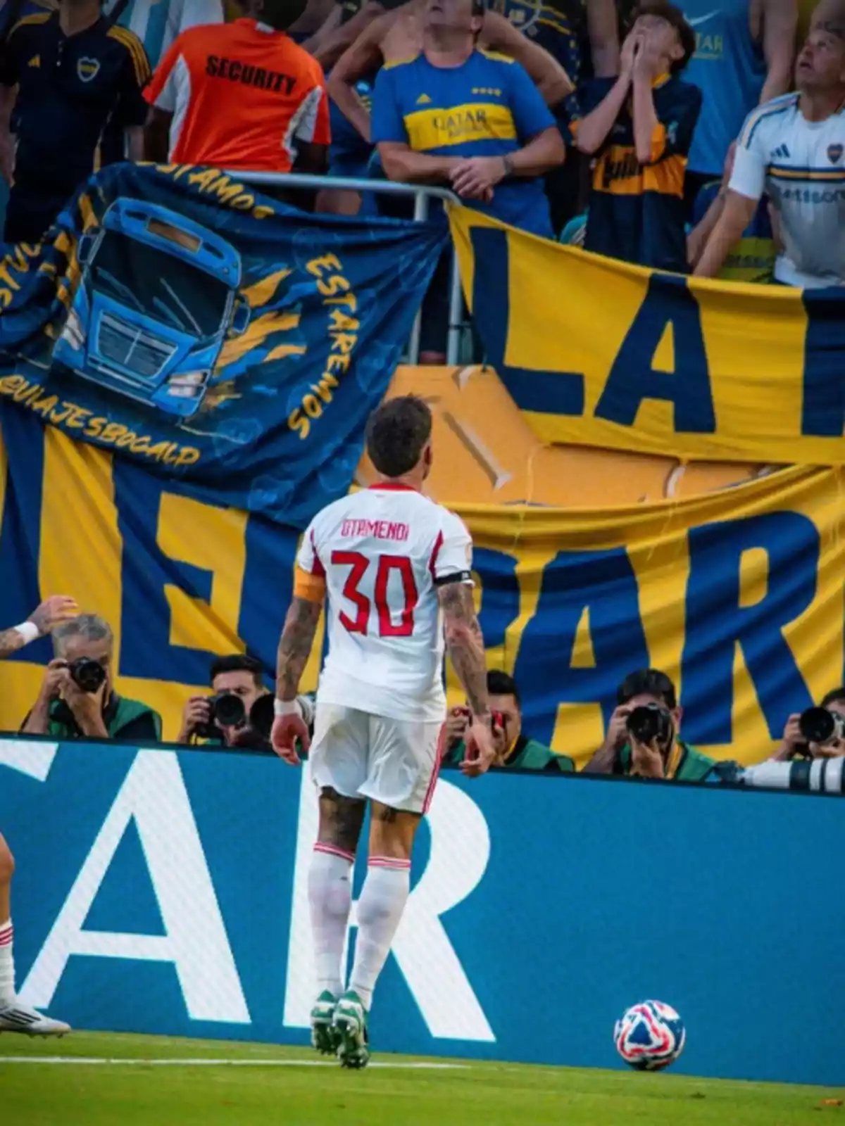 Un futbolista con el número 30 en la camiseta observa a la hinchada rival que sostiene banderas amarillas y azules mientras varios fotógrafos capturan el momento desde el borde del campo. Un futbolista con el número 30 en la camiseta observa a la hinchada rival que sostiene banderas amarillas y azules mientras varios fotógrafos capturan el momento desde el borde del campo.