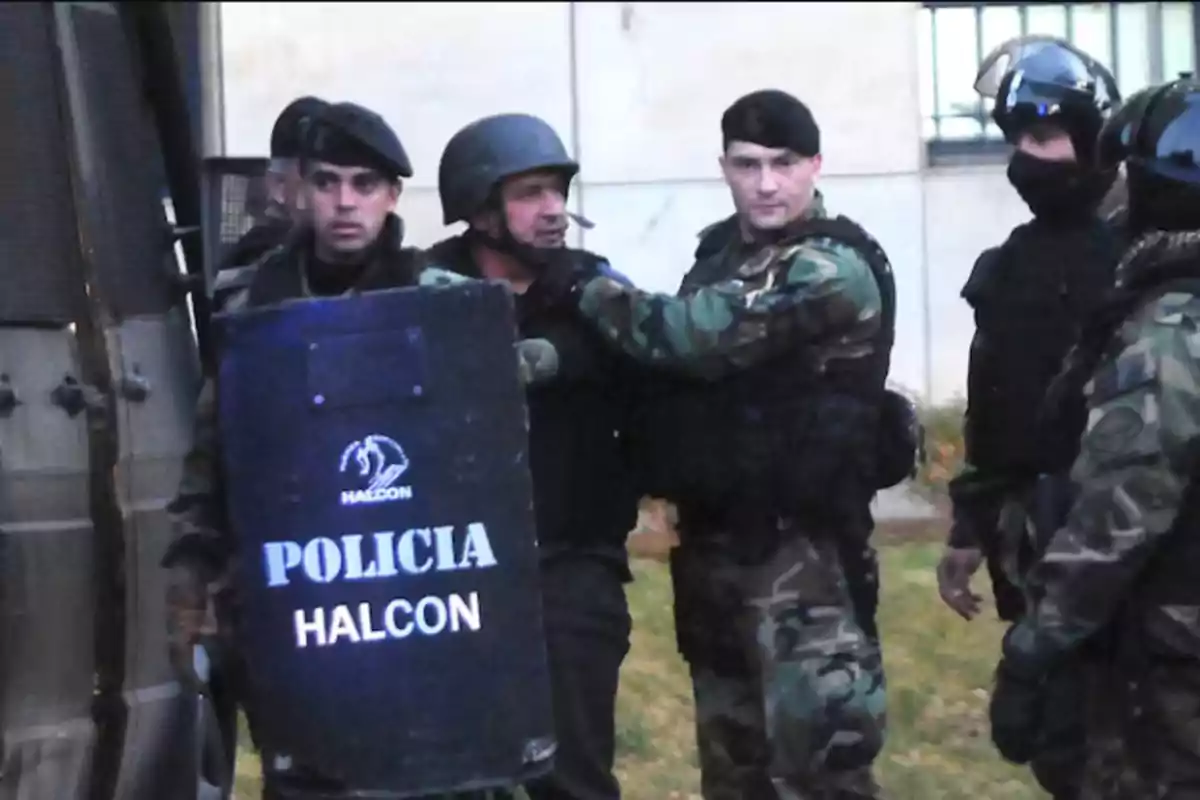 Several heavily armed police officers equipped with shields and bulletproof vests are gathered next to a police vehicle during a tactical operation.