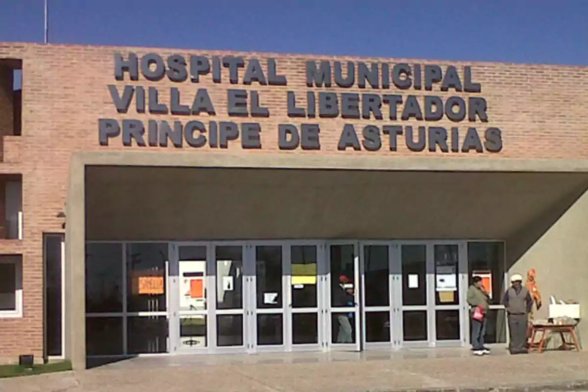 Fachada de un hospital municipal de ladrillo con letras grandes en la parte superior y dos personas de pie cerca de la entrada principal.