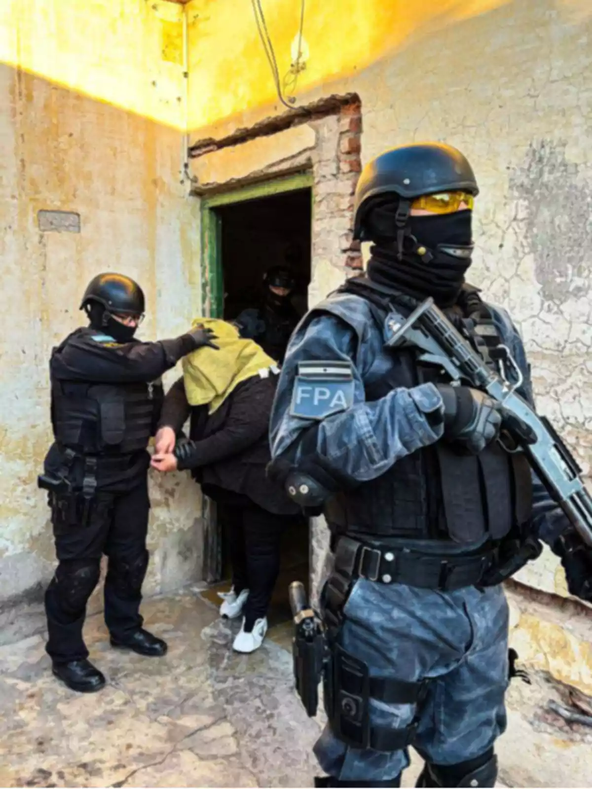 Policías fuertemente armados detienen a una persona con la cabeza cubierta en la entrada de una casa de paredes desgastadas. Policías fuertemente armados detienen a una persona con la cabeza cubierta en la entrada de una casa de paredes desgastadas.