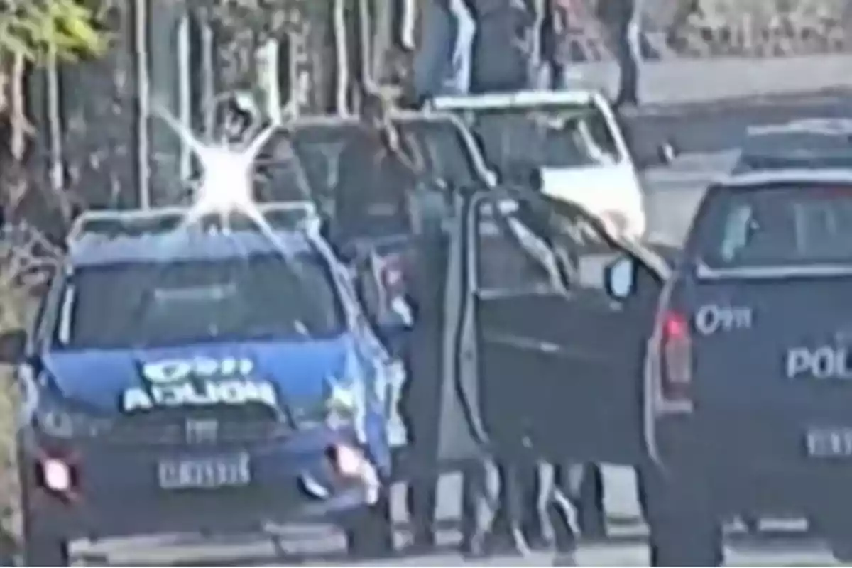 Varias patrullas de policía están detenidas en una calle mientras algunos oficiales se encuentran de pie junto a los vehículos.