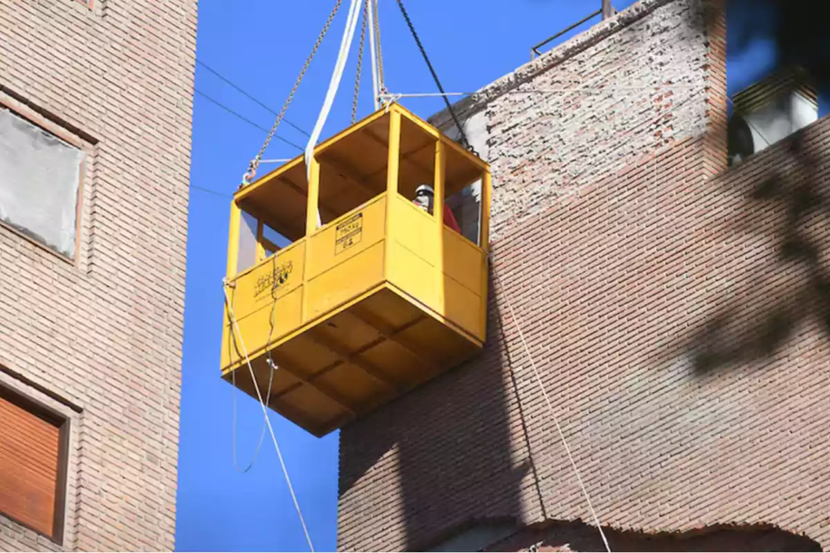 Persona con casco dentro de una plataforma amarilla suspendida por una grúa junto a un edificio de ladrillos