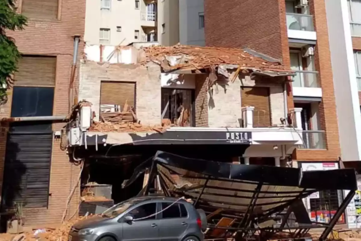 Edificio de dos pisos con el techo y la fachada parcialmente derrumbados, escombros sobre un auto gris y una estructura metálica caída frente al inmueble Edificio de dos pisos con el techo y la fachada parcialmente derrumbados, escombros sobre un auto gris y una estructura metálica caída frente al inmueble