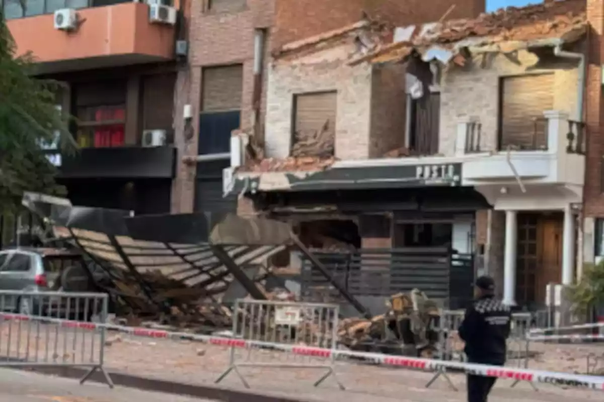 Fachada de edificio con daños severos y escombros en la acera, un auto afectado y una persona observando detrás de una cinta de seguridad Fachada de edificio con daños severos y escombros en la acera, un auto afectado y una persona observando detrás de una cinta de seguridad