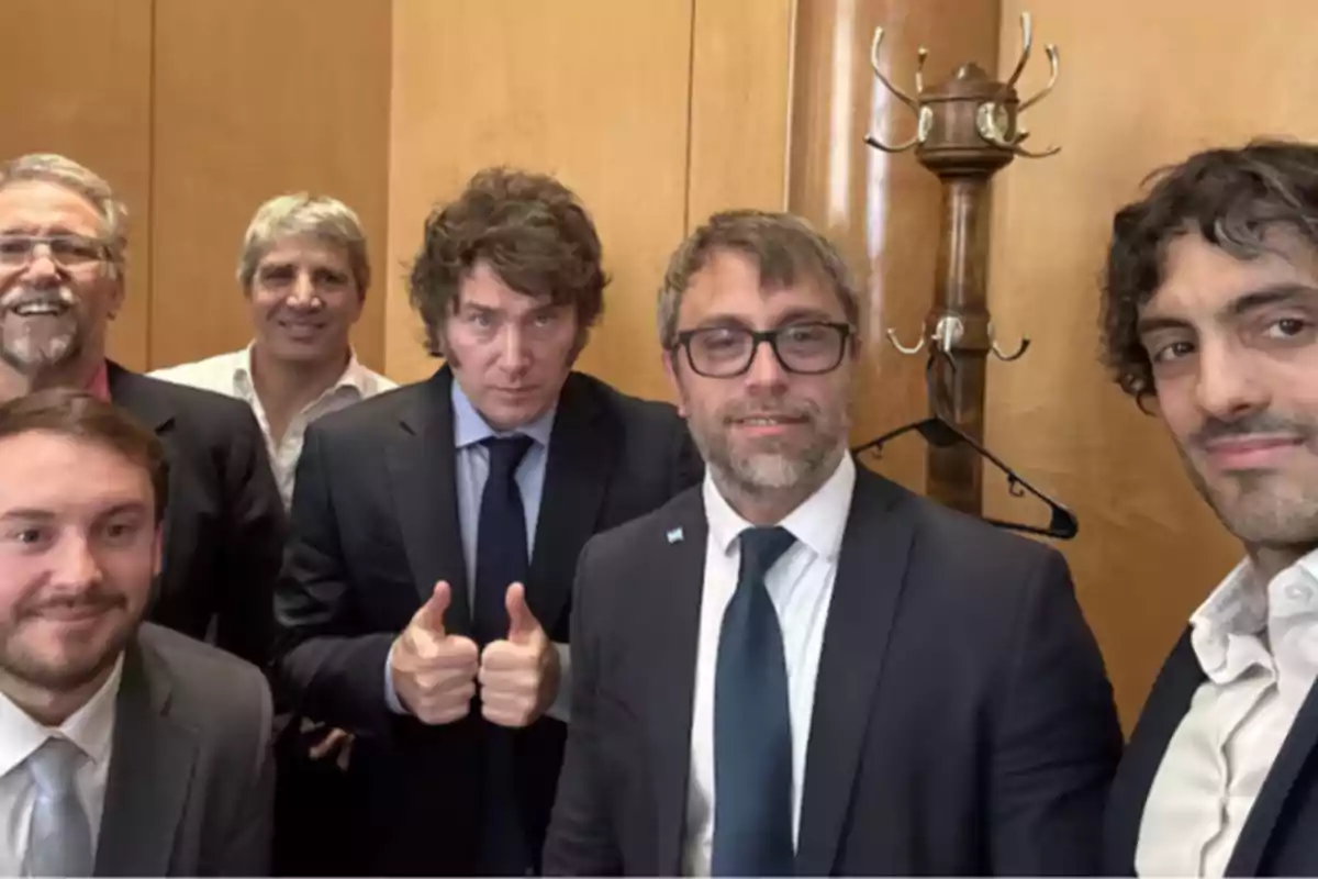 Six men in suits pose for a selfie in an office with wooden walls, one of them giving a thumbs-up gesture.
