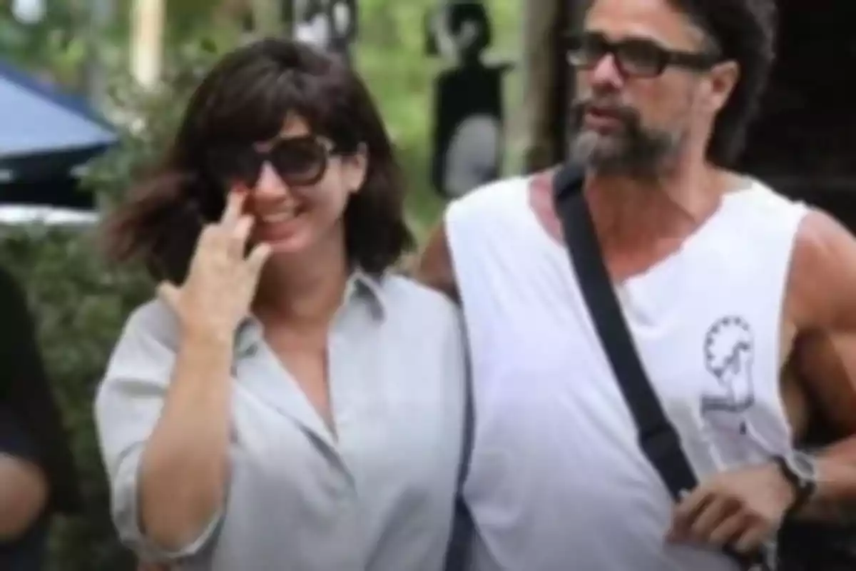 A couple walks outdoors arm in arm, both wearing sunglasses and looking smiling. A couple walks outdoors arm in arm, both wearing sunglasses and looking smiling.