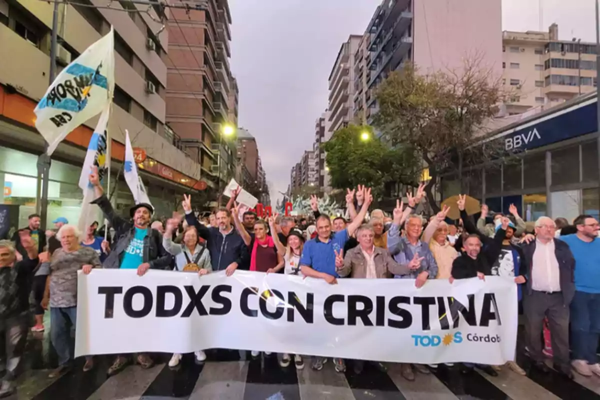 Un grupo de personas marcha por una avenida de la ciudad sosteniendo una pancarta que dice "TODXS CON CRISTINA" mientras hacen el gesto de la victoria con las manos y llevan banderas.