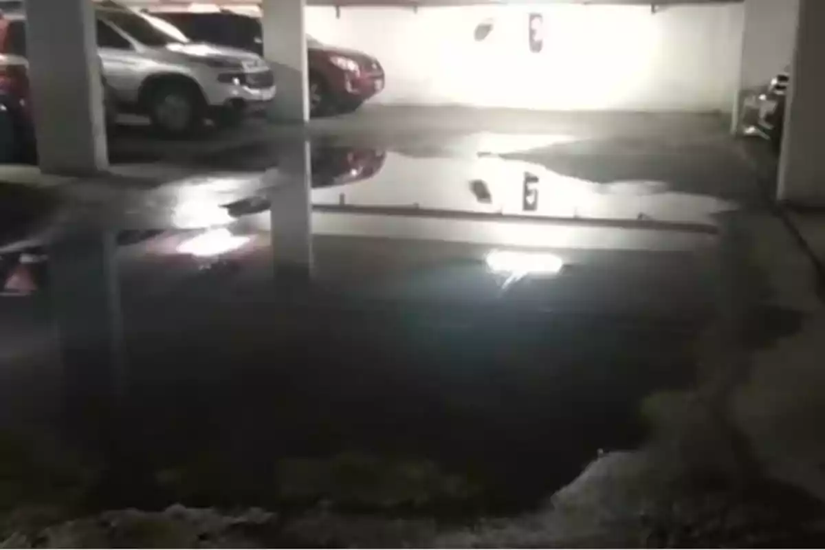 Estacionamiento subterráneo con charco de agua reflejando la luz y los autos estacionados.
