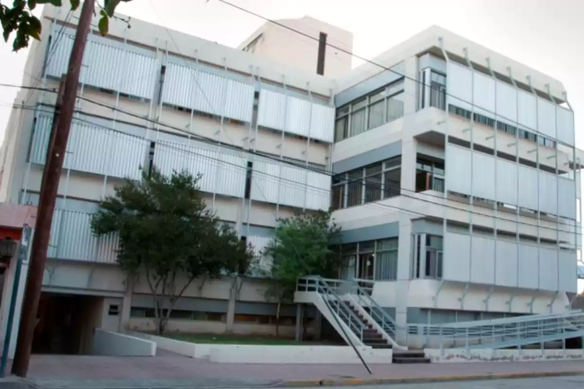 Edificio moderno de varios pisos con ventanas grandes y persianas metálicas en la fachada rodeado de árboles y una rampa de acceso en la entrada