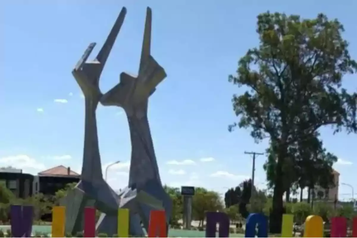 Escultura de dos manos entrelazadas en una plaza con letras de colores y un árbol al fondo en un día soleado Escultura de dos manos entrelazadas en una plaza con letras de colores y un árbol al fondo en un día soleado