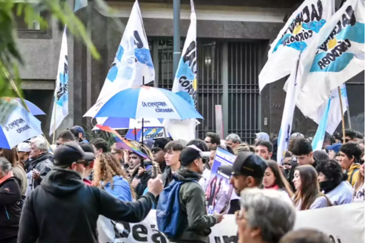 Manifestación con personas sosteniendo pancartas, banderas y paraguas azules y blancos en una calle frente a un edificio