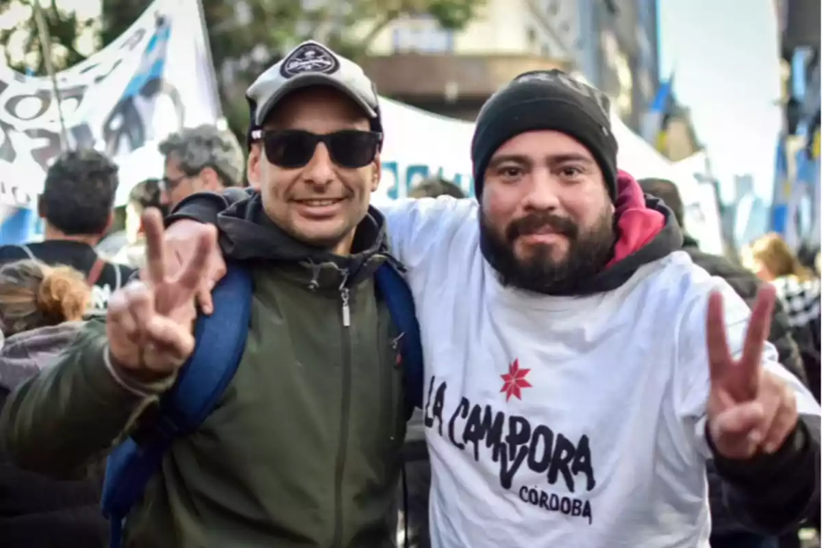 Dos personas sonríen y hacen el gesto de la victoria con los dedos mientras participan en una manifestación, una de ellas lleva una camiseta blanca con la inscripción La Cámpora Córdoba. Dos personas sonríen y hacen el gesto de la victoria con los dedos mientras participan en una manifestación, una de ellas lleva una camiseta blanca con la inscripción La Cámpora Córdoba.