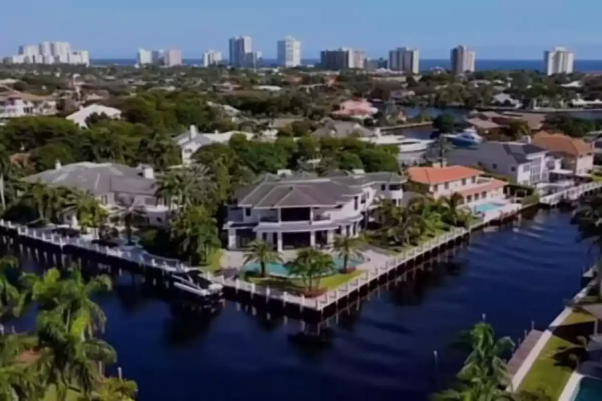 Vista aérea de una lujosa casa junto a un canal con piscina y palmeras en una zona residencial, al fondo se observan edificios altos y el mar Vista aérea de una lujosa casa junto a un canal con piscina y palmeras en una zona residencial, al fondo se observan edificios altos y el mar