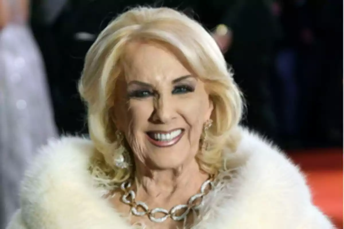 Smiling older woman with blonde hair, elegantly dressed in a white coat and a silver necklace