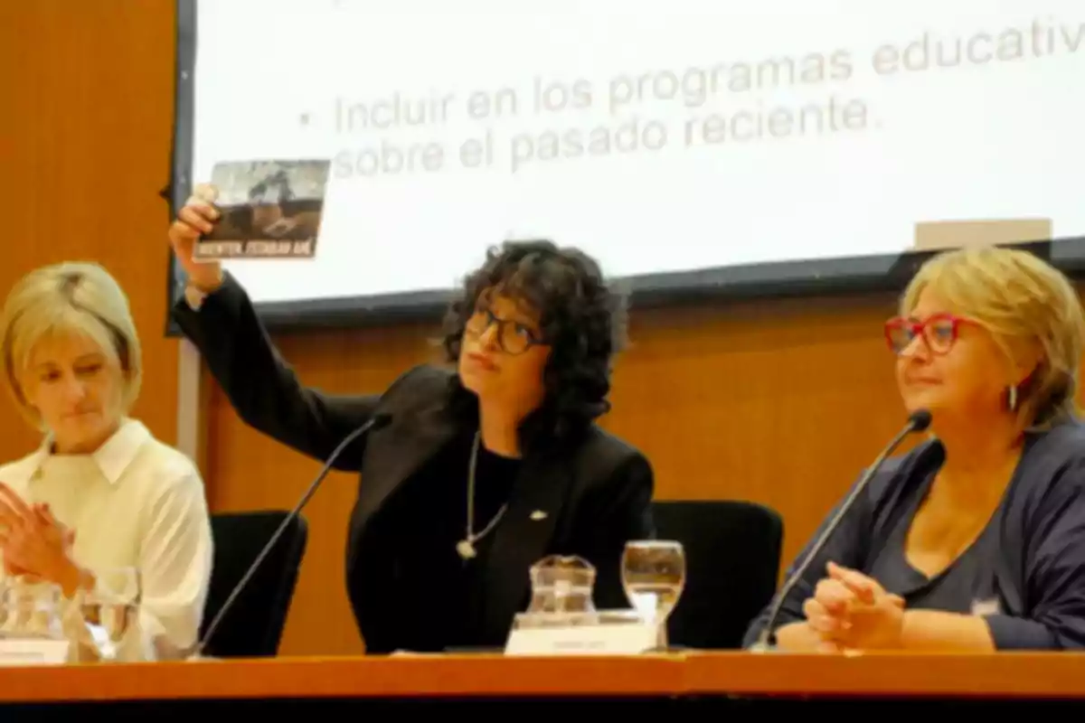 Three people are sitting at a table during a conference, one of them is holding a book while the other two watch.