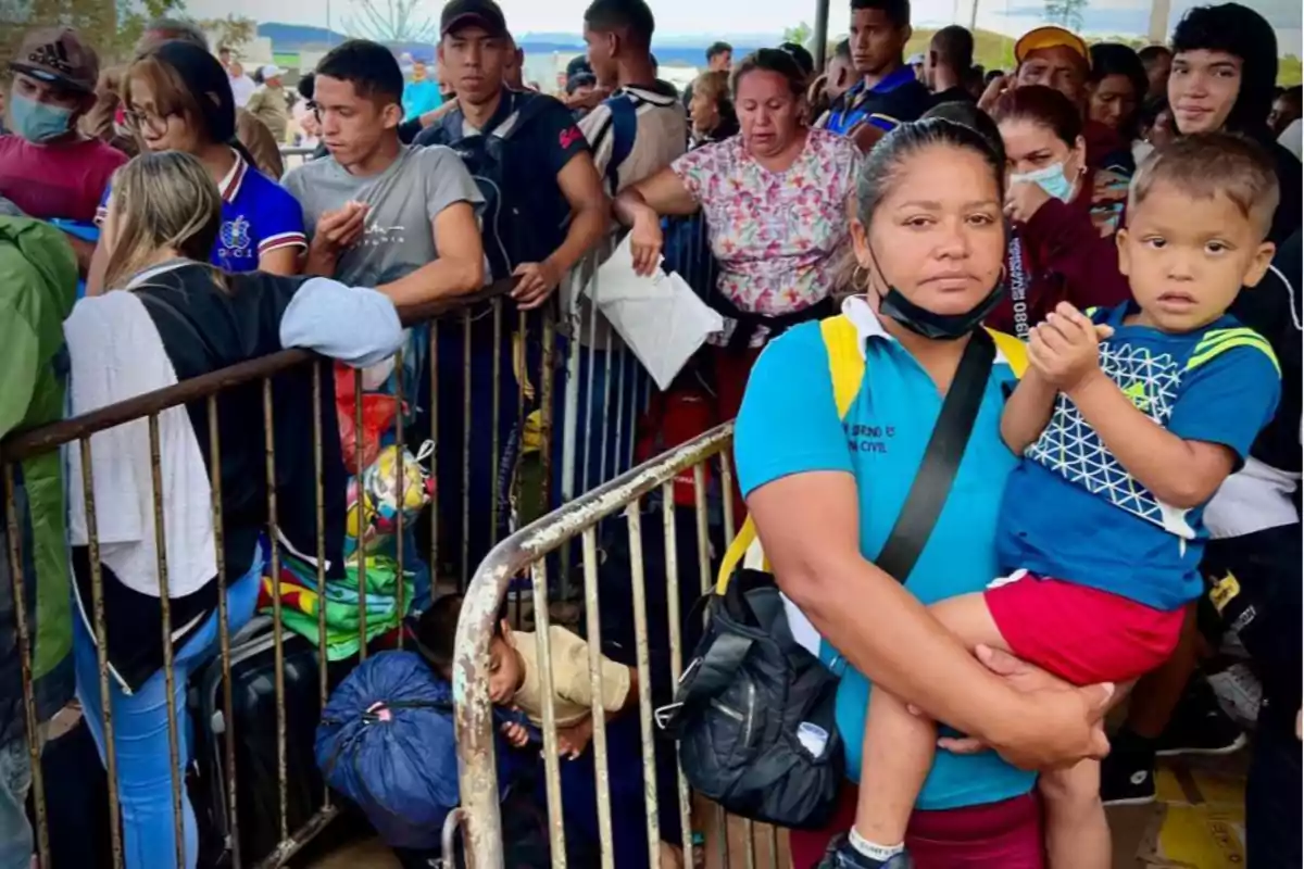 A group of people waits in line behind a metal fence; a woman at the front is holding a small child in her arms, both looking at the camera, while others watch or talk to each other, some carrying backpacks and bags.