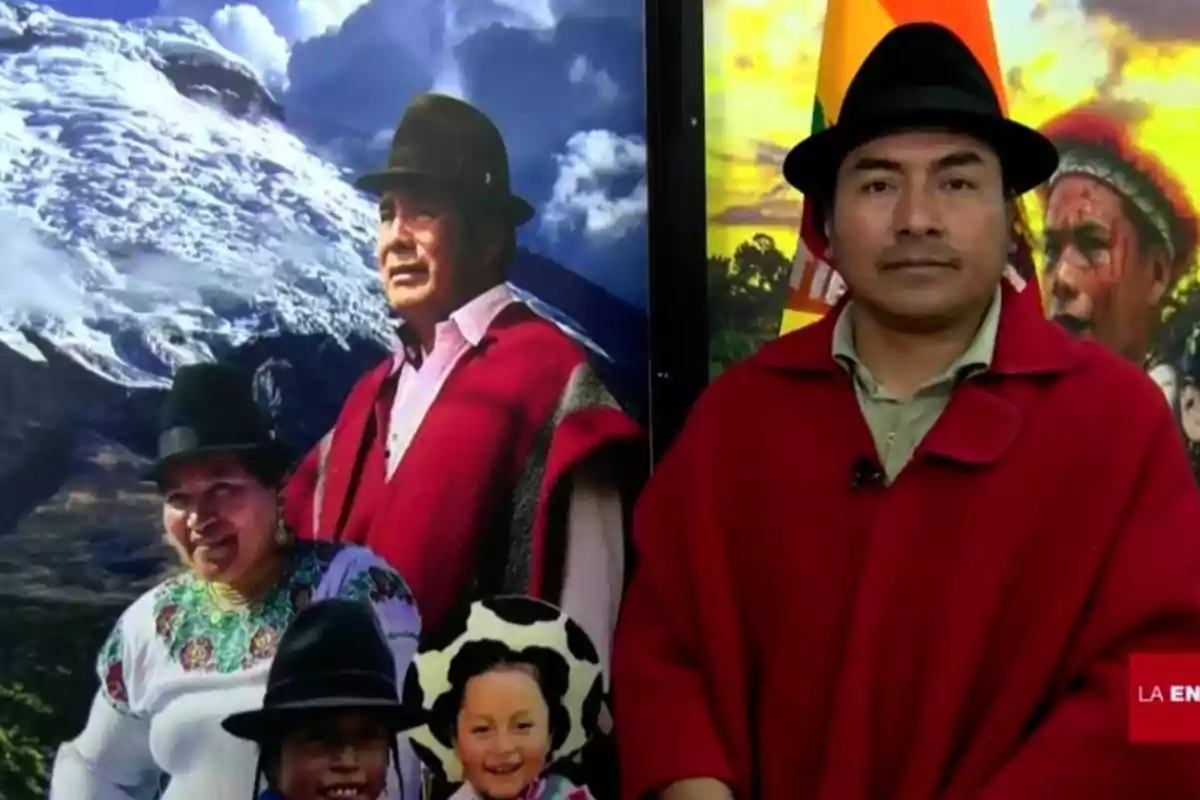 Un hombre con sombrero y poncho rojo está de pie junto a un cartel que muestra a una familia con vestimenta tradicional indígena frente a un paisaje montañoso nevado.