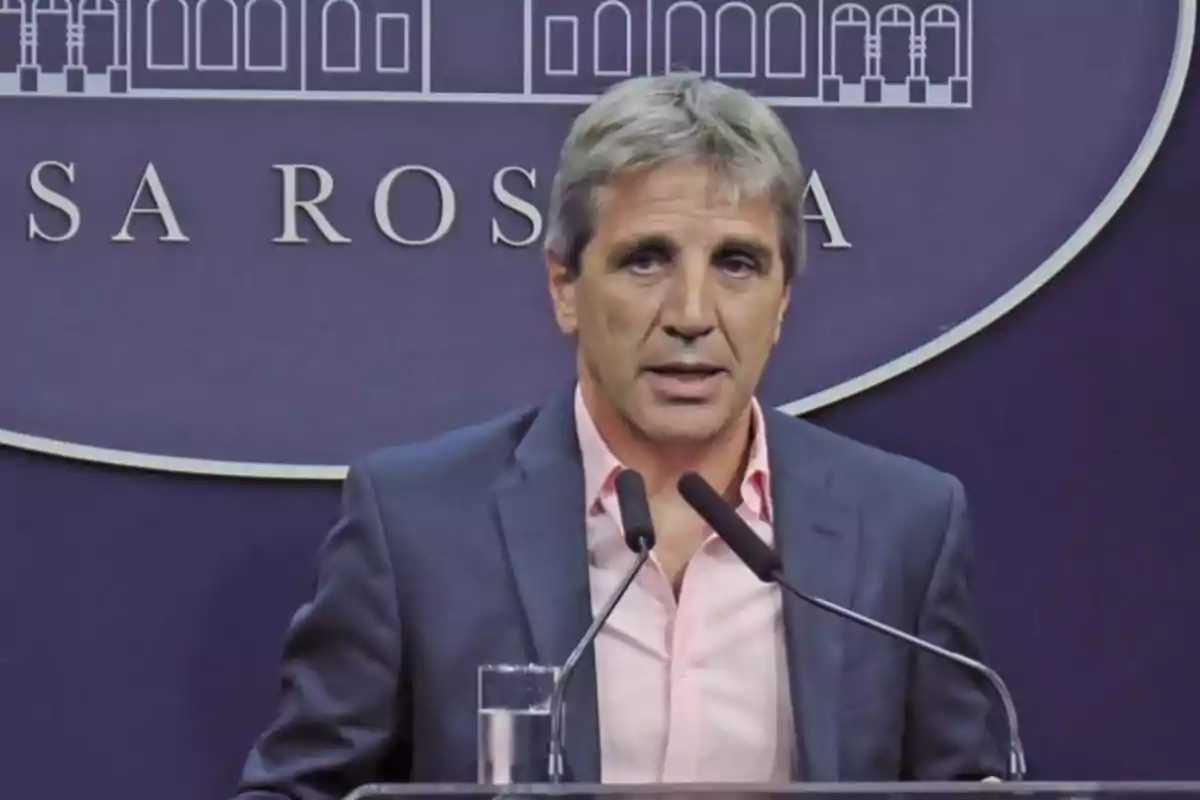 A gray-haired man in a dark suit and pink jersey speaks in front of two microphones with a background featuring letters and an architectural design.