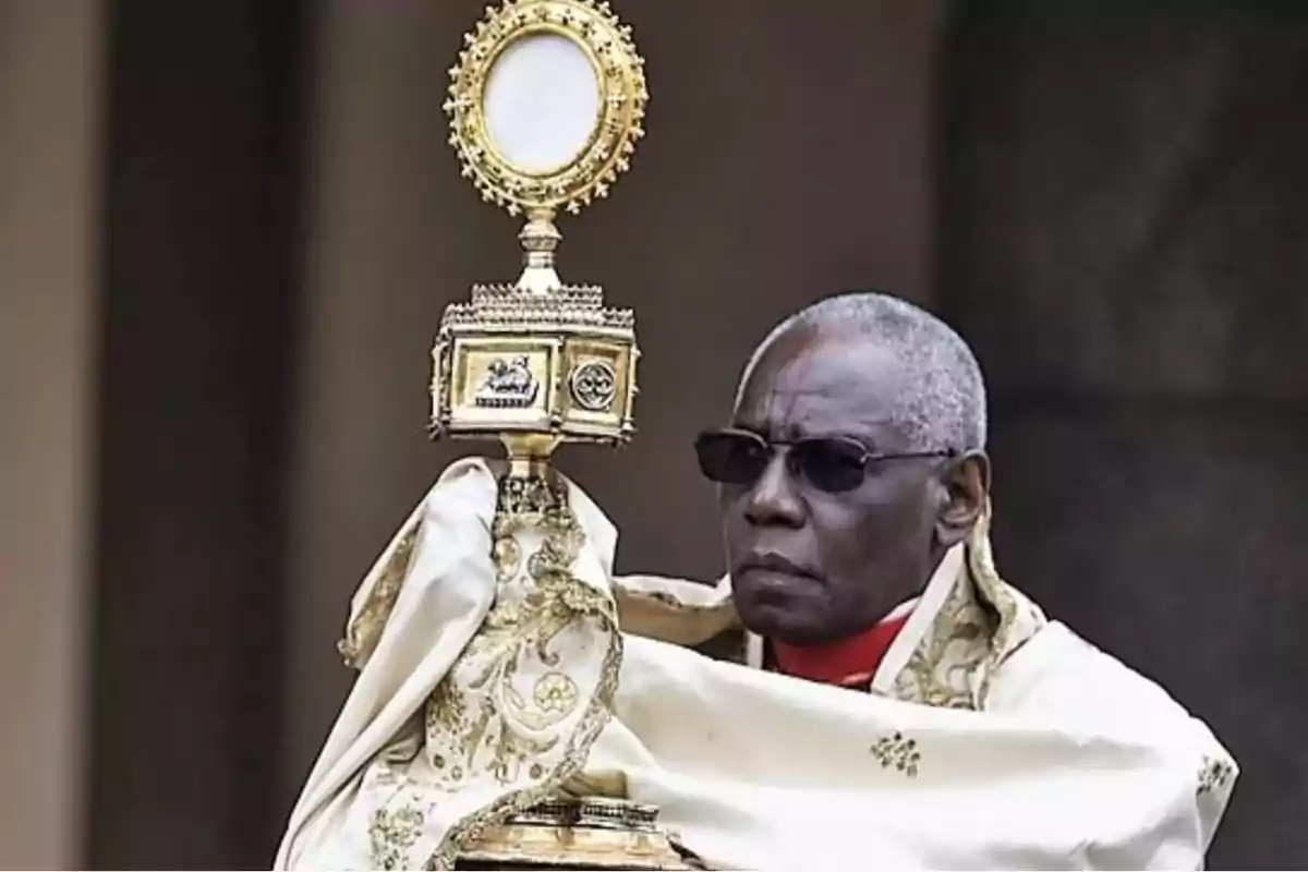 A man with sunglasses holds a golden monstrance decorated with religious details while wearing a white and gold ceremonial cape.