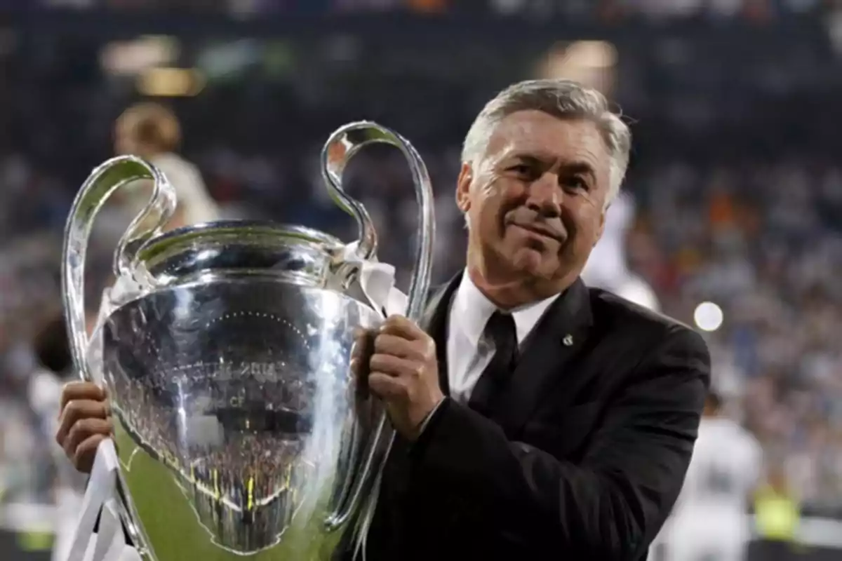 Un hombre de traje sostiene un gran trofeo plateado en un estadio lleno de espectadores. Un hombre de traje sostiene un gran trofeo plateado en un estadio lleno de espectadores.