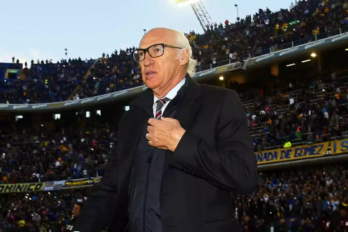 A man with glasses and a dark suit walks in a stadium full of spectators.