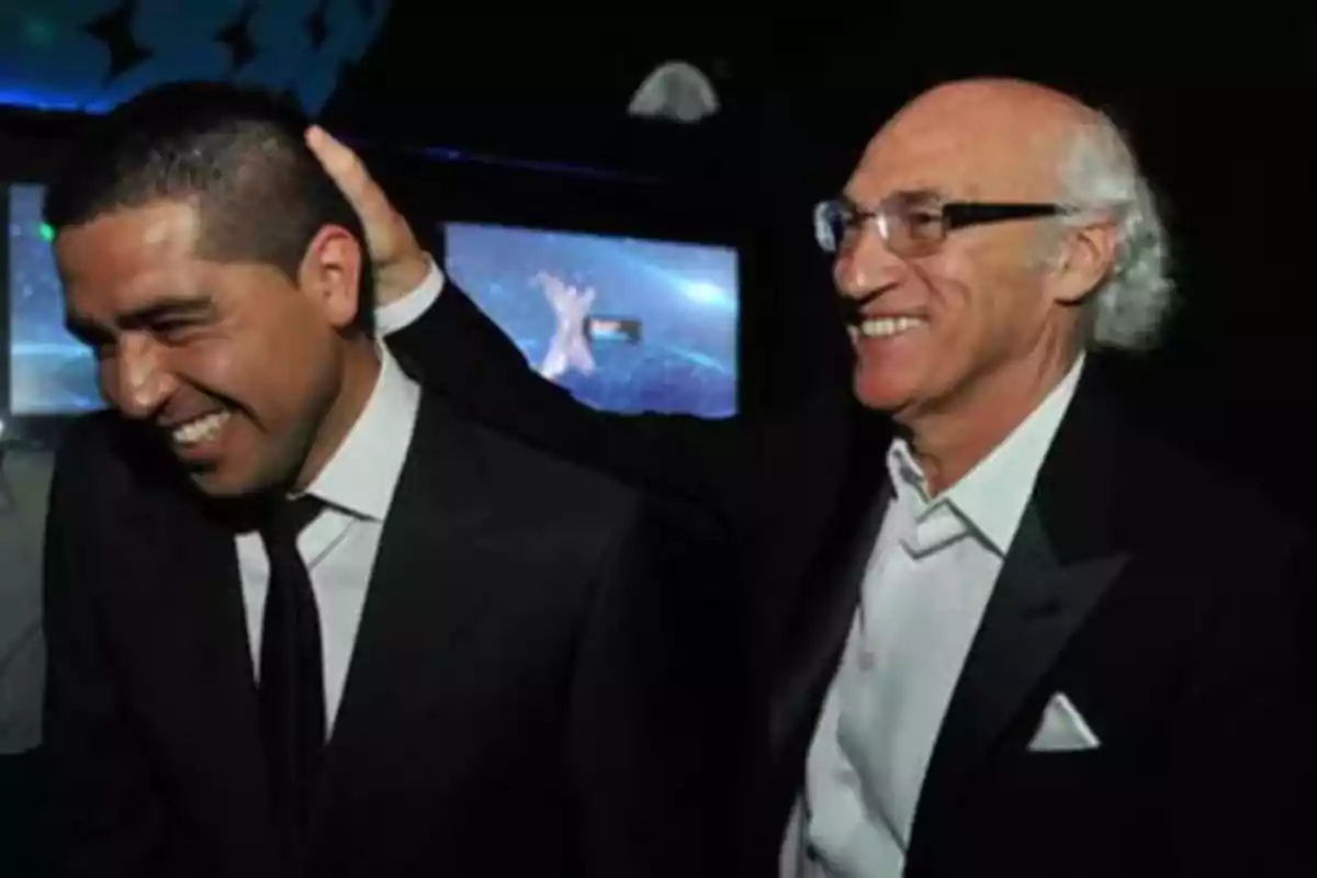 Dos hombres sonríen mientras uno de ellos toca la cabeza del otro en un ambiente oscuro y elegante. Dos hombres sonríen mientras uno de ellos toca la cabeza del otro en un ambiente oscuro y elegante.