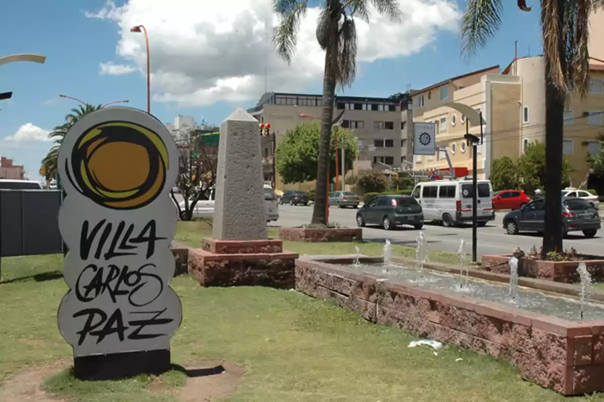 Una señal de bienvenida a Villa Carlos Paz junto a una fuente y un obelisco en un área urbana con palmeras y edificios al fondo.