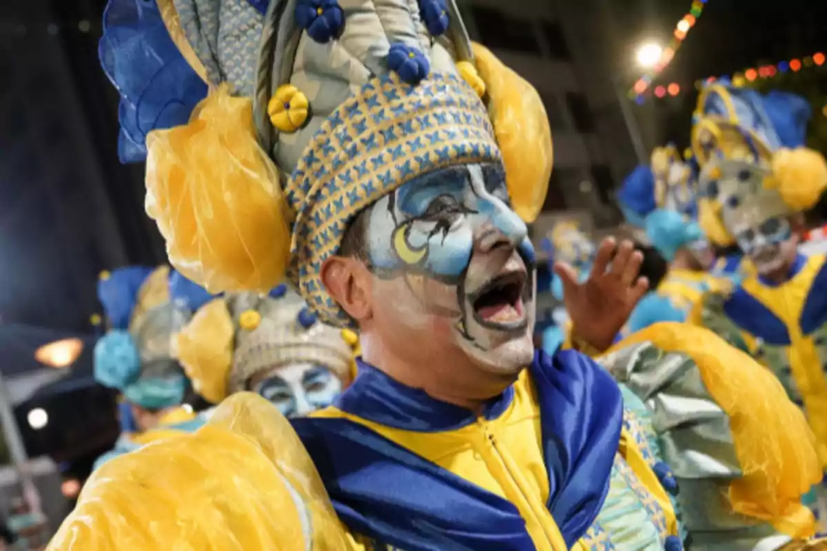 Person dressed in a colorful carnival costume in blue and yellow tones, with artistic makeup on the face and an elaborate decorated hat.