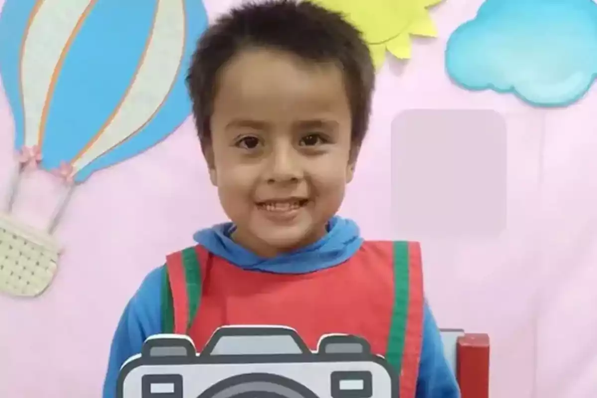 Un niño sonriente con un delantal rojo sostiene una figura de cámara frente a un fondo decorado con un globo aerostático y nubes. Un niño sonriente con un delantal rojo sostiene una figura de cámara frente a un fondo decorado con un globo aerostático y nubes.