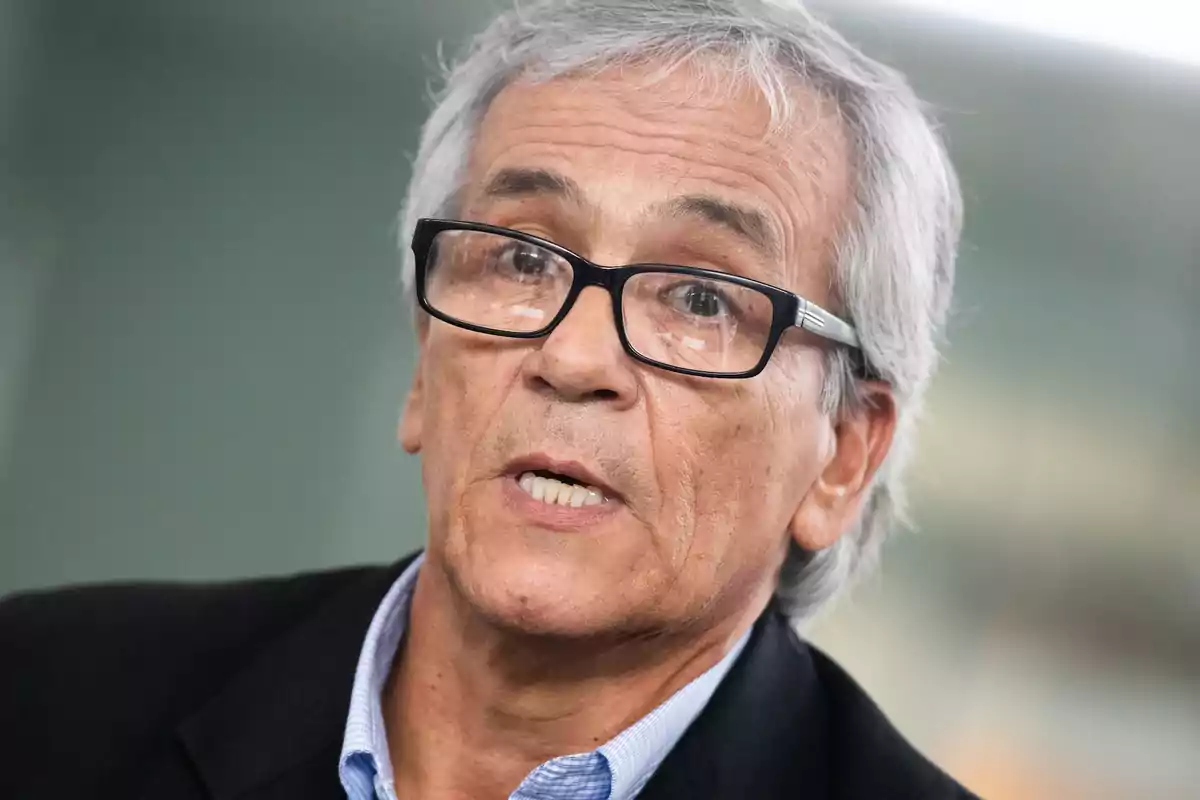 Hombre de cabello canoso y gafas negras hablando con expresión seria