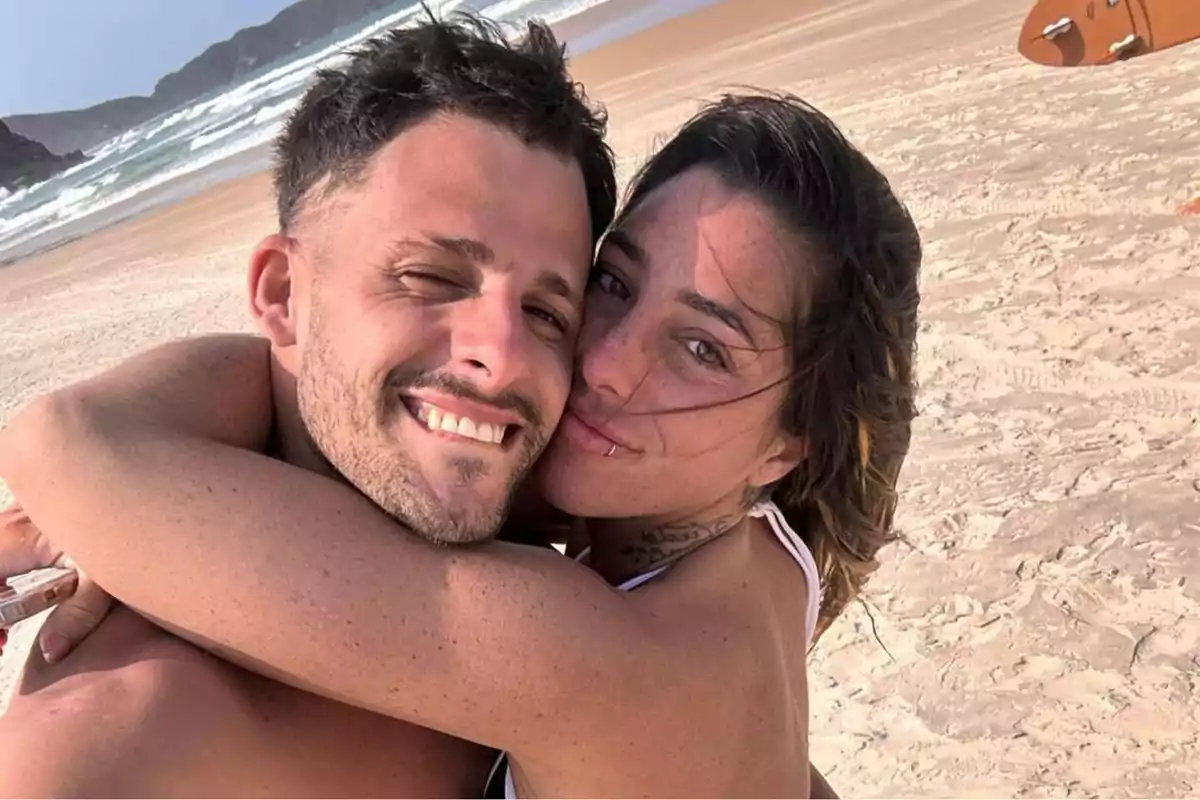 Una pareja sonriente se abraza en la playa con el mar y las olas de fondo.