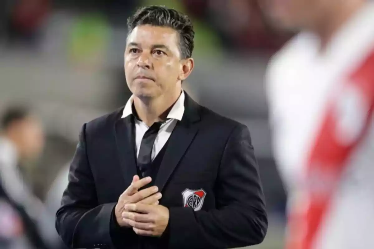 Un hombre con traje oscuro y corbata desajustada, con un escudo de River Plate en el bolsillo, está de pie en un estadio.