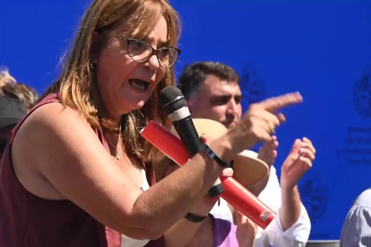A woman with glasses and brown hair holds a microphone while gesturing passionately at an outdoor event. A woman with glasses and brown hair holds a microphone while gesturing passionately at an outdoor event.
