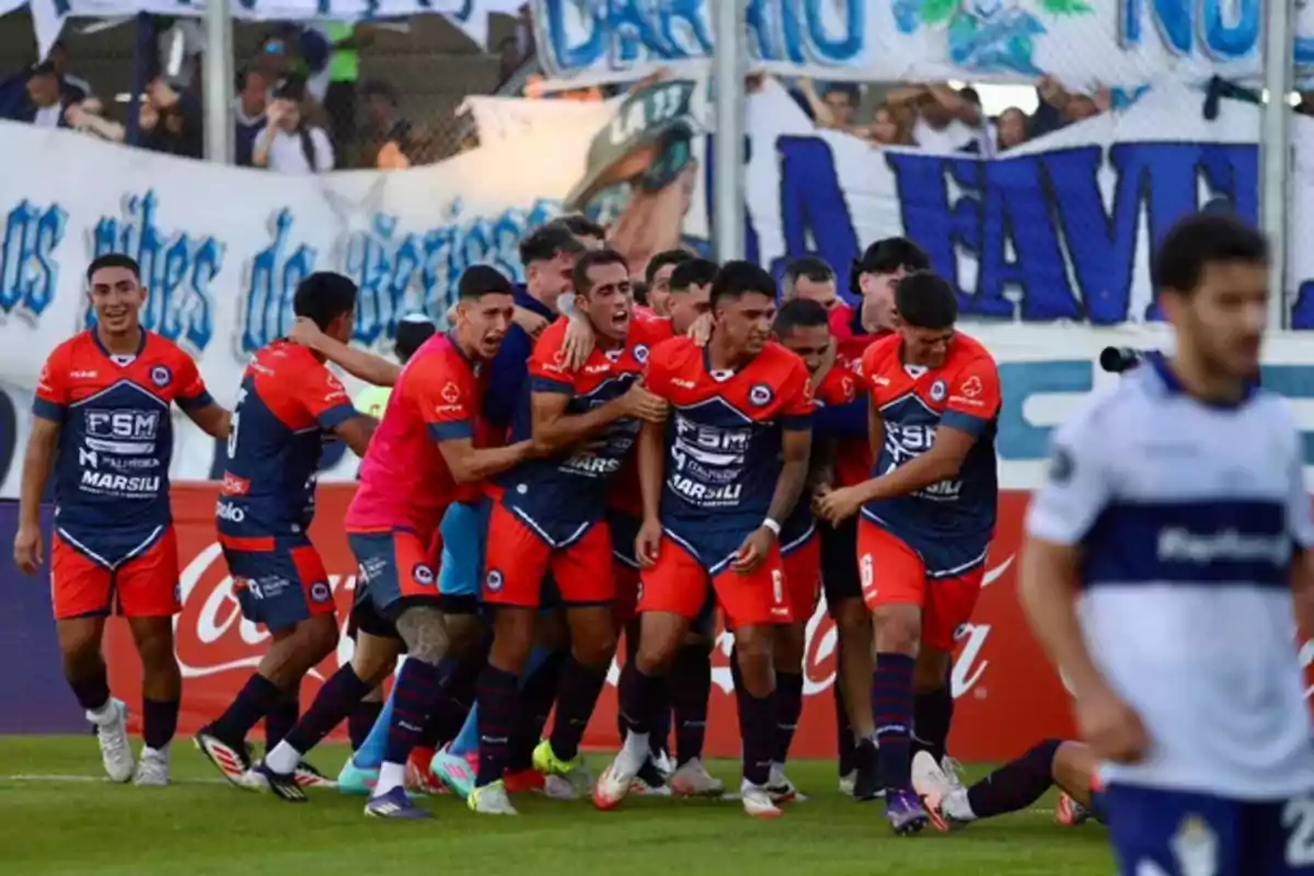 Un grupo de jugadores de fútbol con uniformes rojos y azules celebra en el campo mientras un jugador del equipo contrario camina cerca. Un grupo de jugadores de fútbol con uniformes rojos y azules celebra en el campo mientras un jugador del equipo contrario camina cerca.