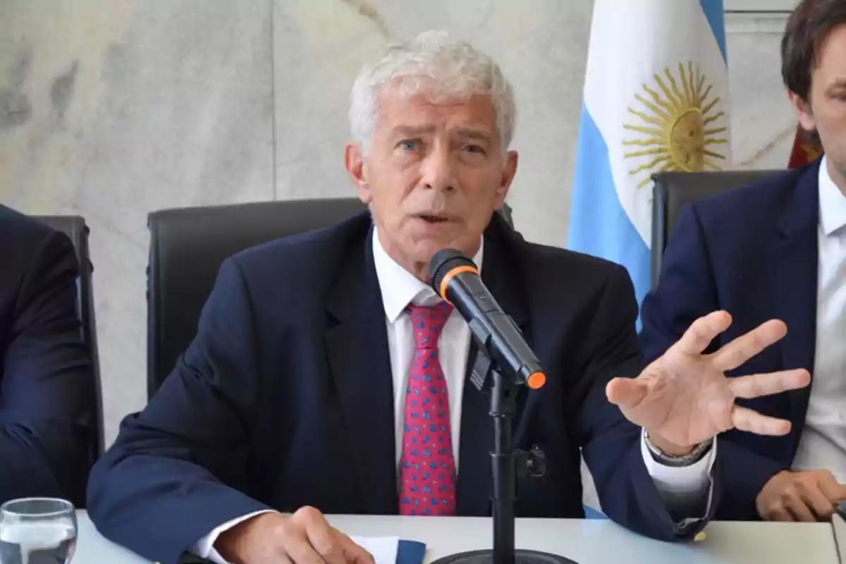 Un hombre de cabello canoso y traje oscuro habla en un micrófono durante una conferencia de prensa con una bandera argentina de fondo.