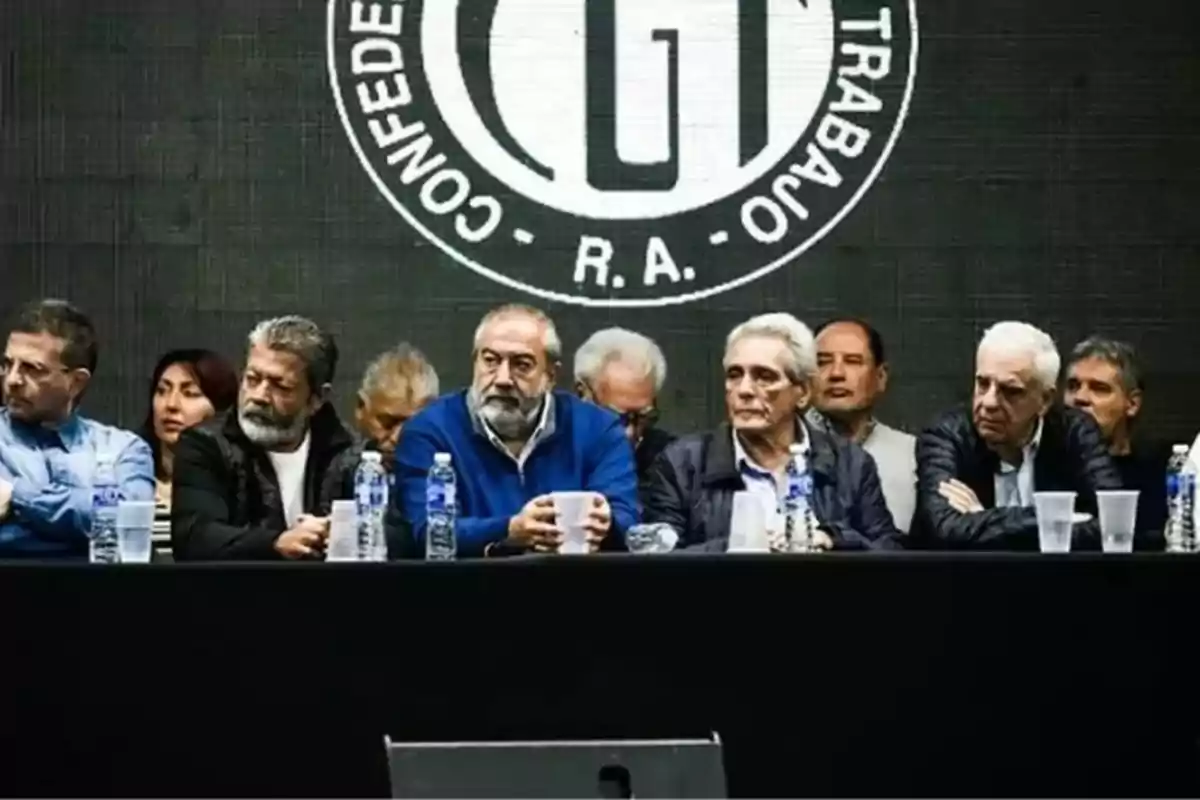 Un grupo de personas sentadas en una mesa con botellas de agua y vasos, frente a un fondo con un logotipo circular que dice 