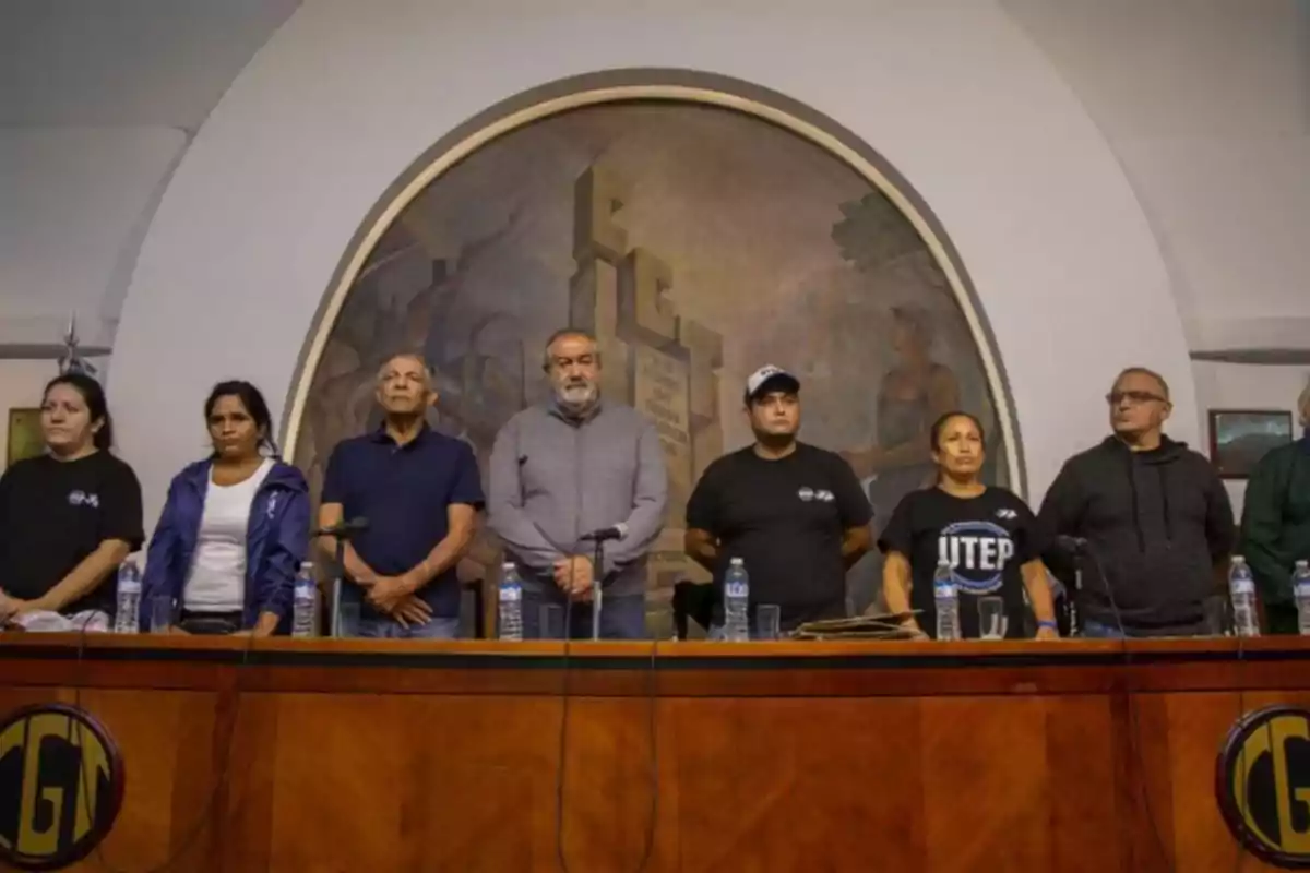 Un grupo de personas está de pie detrás de una mesa en una sala con un mural en la pared.