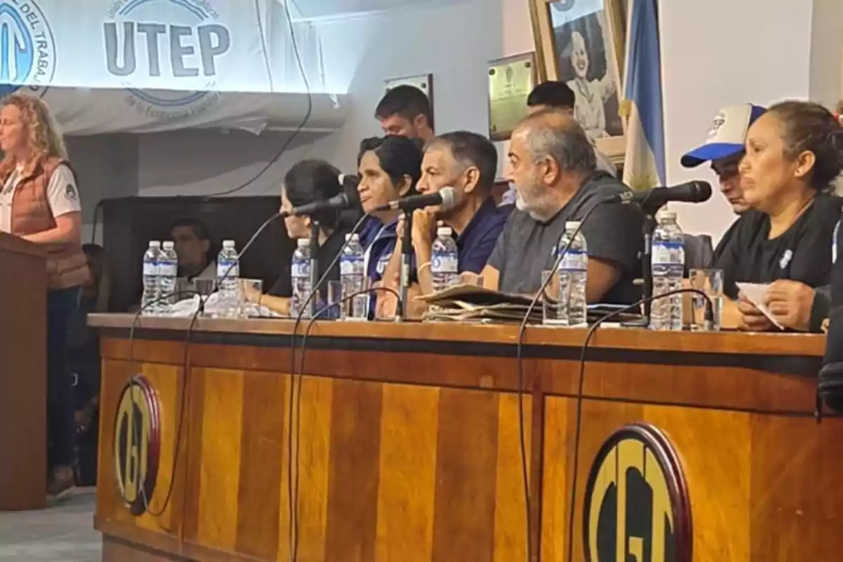 Un grupo de personas está sentado en una mesa con micrófonos y botellas de agua, mientras una mujer de pie habla en un podio, en un entorno que parece ser una conferencia o reunión formal, con un cartel de UTEP en el fondo.