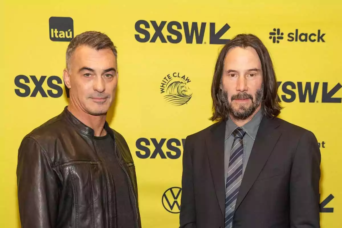 Two men pose in front of a yellow background with SXSW logos and various brands; one is wearing a black leather jacket and the other is dressed in a dark suit with a tie. Two men pose in front of a yellow background with SXSW logos and various brands; one is wearing a black leather jacket and the other is dressed in a dark suit with a tie.