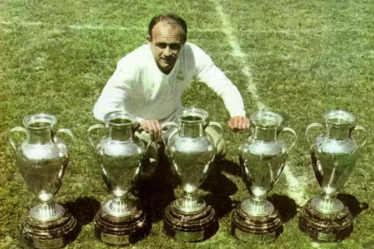 Un hombre en un campo de fútbol posando con cinco trofeos idénticos.