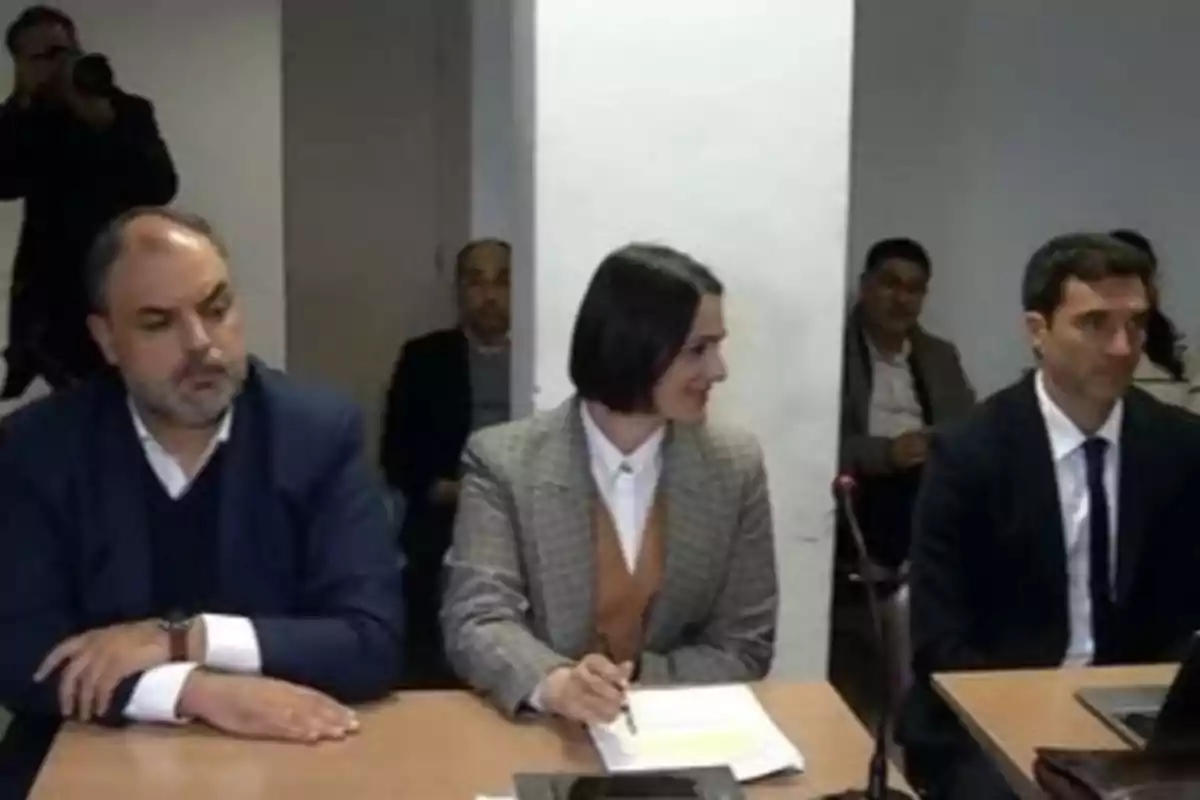 Three people in formal attire are seated at a table during a meeting or hearing, while others watch in the background. Three people in formal attire are seated at a table during a meeting or hearing, while others watch in the background.